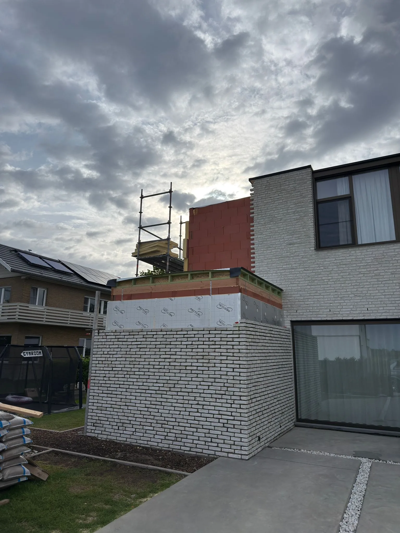 Uitbreiding in aanbouw achter een woning met witte gevelsteen, met isolatieplaten, stelling en een houten dakrand