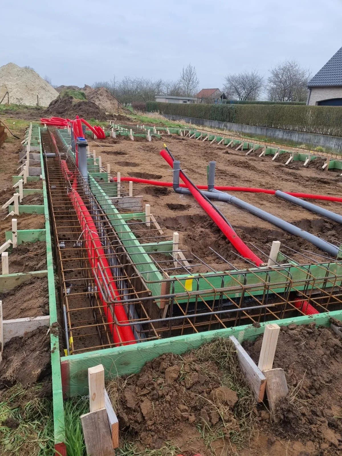 Funderingswerf van een nieuwbouw met wapening in de sleuven, groene bekisting, rode mantelbuizen en grijze afvoerbuizen