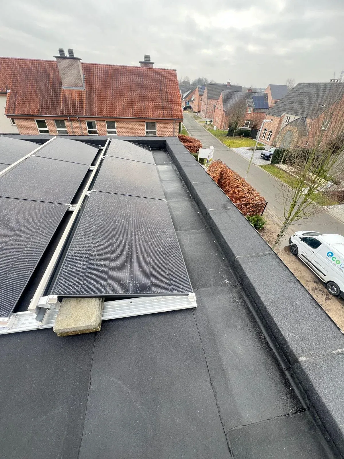 Rij zwarte zonnepanelen op een plat dak met zicht op een woonstraat met bakstenen huizen